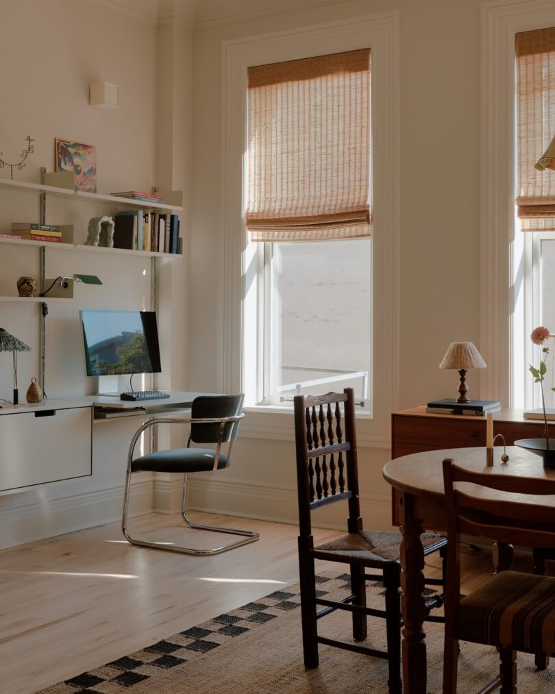 Oficina soleada en casa con escritorio montado en la pared, computadora, silla negra, estantería, mesa de comedor redonda, sillas de madera y dos grandes ventanales con persianas tejidas.