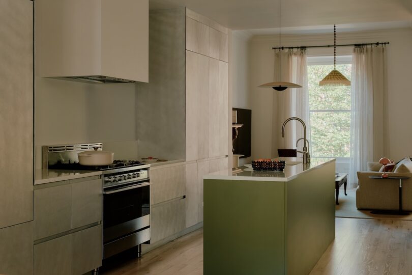 Cocina moderna con gabinetes mate, estufa a gas, isla con fregadero y sala de estar con grandes ventanales y cortinas transparentes.