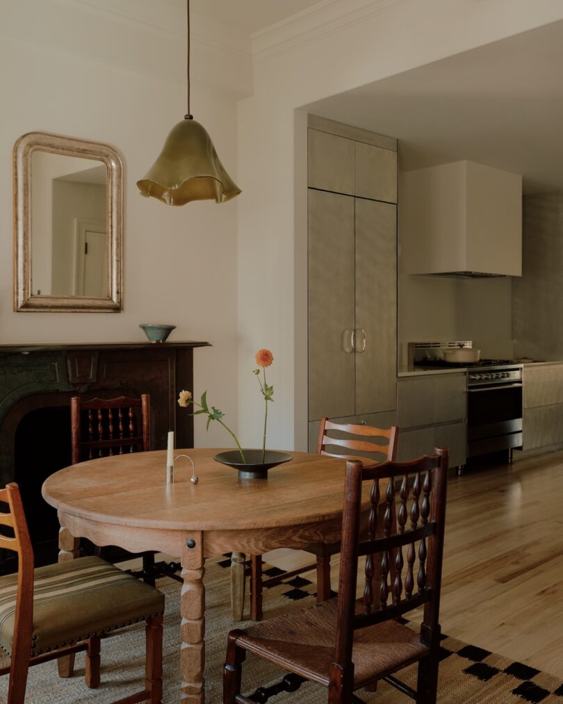 La cocina minimalista cuenta con una mesa de comedor redonda de madera con tres sillas, pisos de madera clara, luces colgantes, espejos y arreglos florales sencillos.