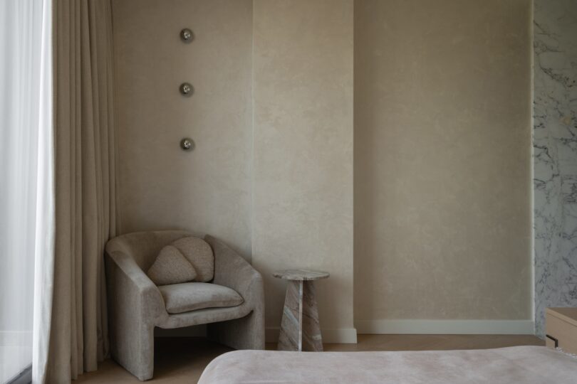 Esquina de dormitorio minimalista con sillón beige, pequeña mesa auxiliar redonda y tres decoraciones metálicas en una pared de textura clara.