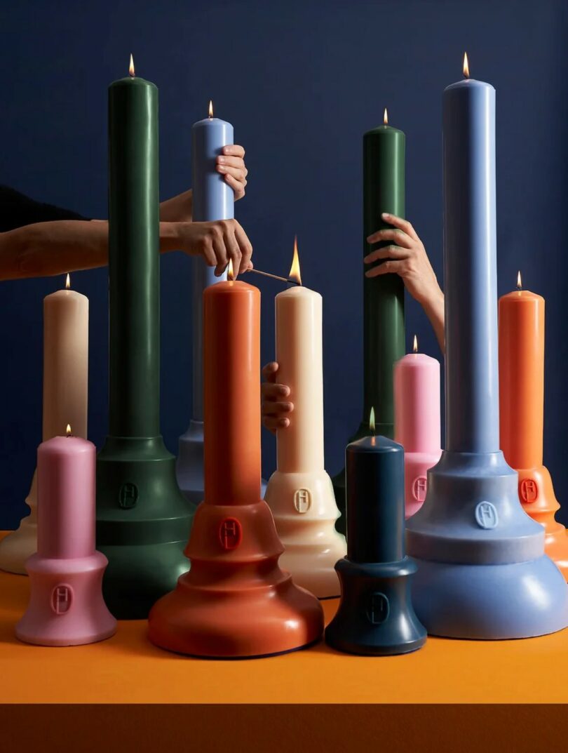 Varias manos encienden velas talladas, altas y cortas, de diferentes colores sobre una superficie naranja sobre un fondo azul oscuro.