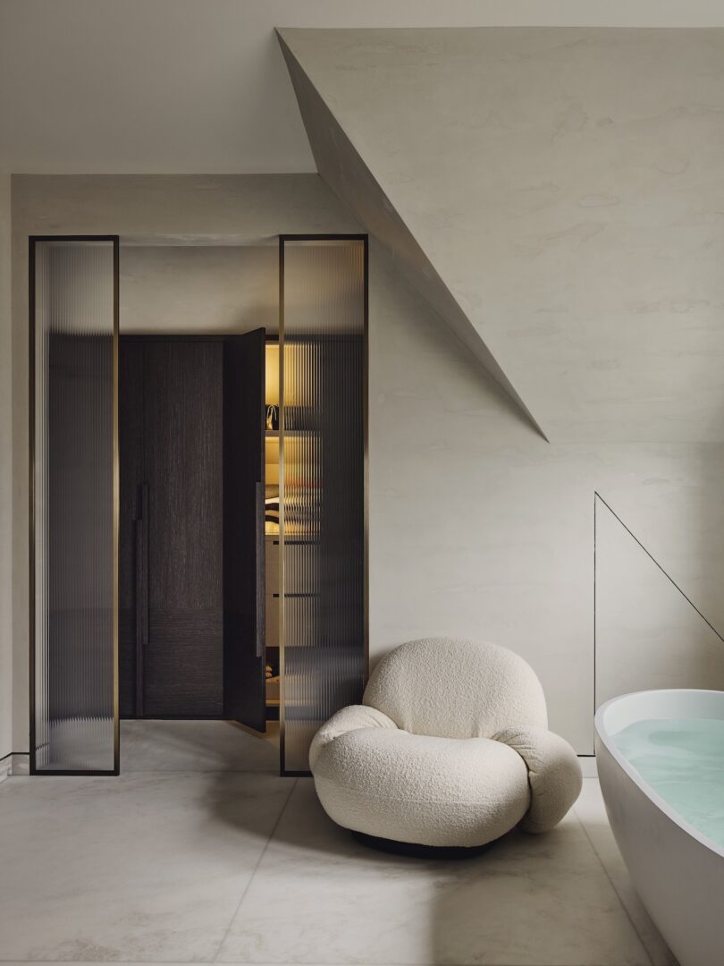 El baño minimalista cuenta con una chaise longue color crema texturizada, puertas dobles de vidrio, almacenamiento incorporado y una bañera blanca parcialmente llena debajo de un techo inclinado.