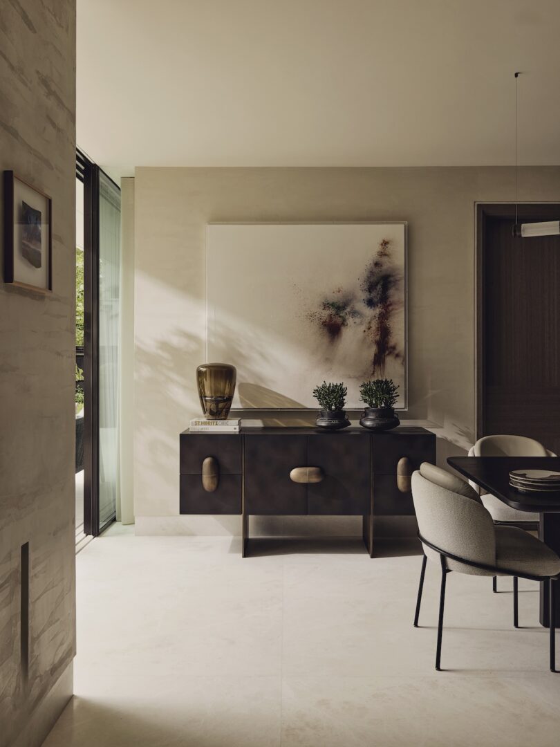 El moderno comedor cuenta con un aparador oscuro, dos plantas en macetas, un jarrón de vidrio marrón y arte abstracto en la pared. La luz natural entra a raudales por los grandes ventanales, iluminando la habitación en tonos neutros.