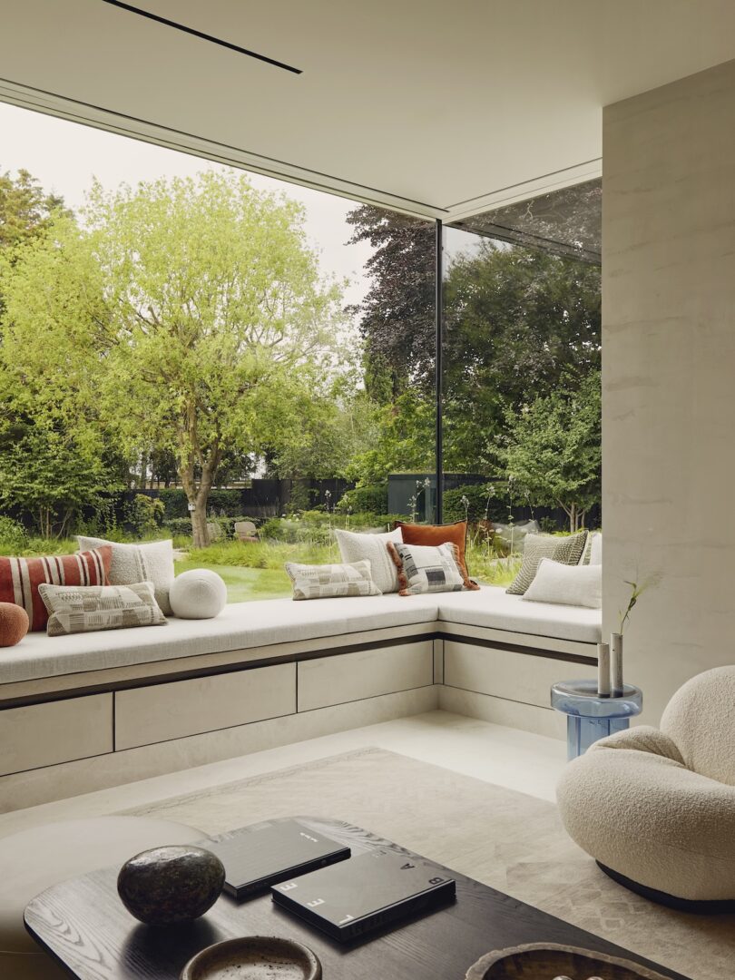 Sala de estar moderna con asiento incorporado junto a la ventana, cojines y un gran ventanal en esquina con vista a un jardín verde, amueblado en tonos neutros y mesa de centro con detalles decorativos.