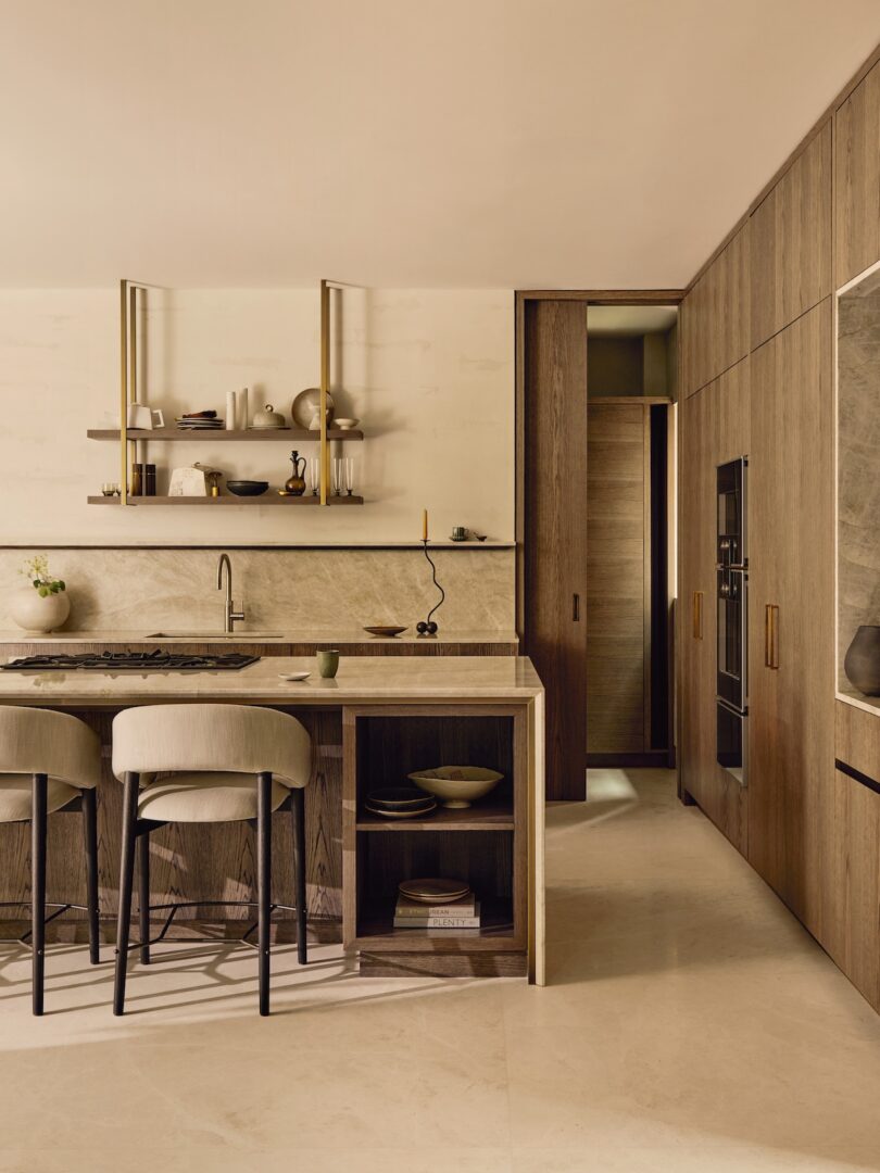 Cocina moderna con gabinetes de madera, encimeras de mármol, estanterías abiertas con vajilla, electrodomésticos integrados y dos taburetes tapizados en la isla.