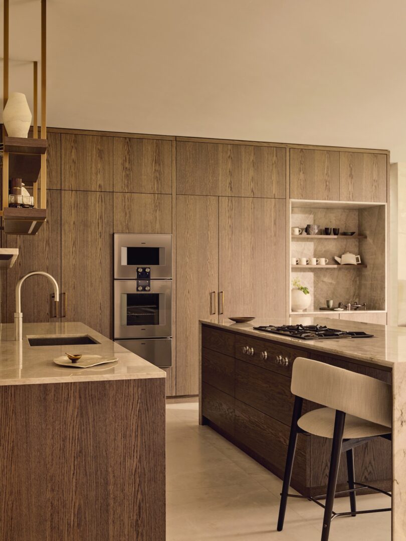 La moderna cocina cuenta con gabinetes de madera, electrodomésticos integrados de acero inoxidable, una isla central con estufa y asientos, y estantes abiertos para exhibir tazas y tazones.