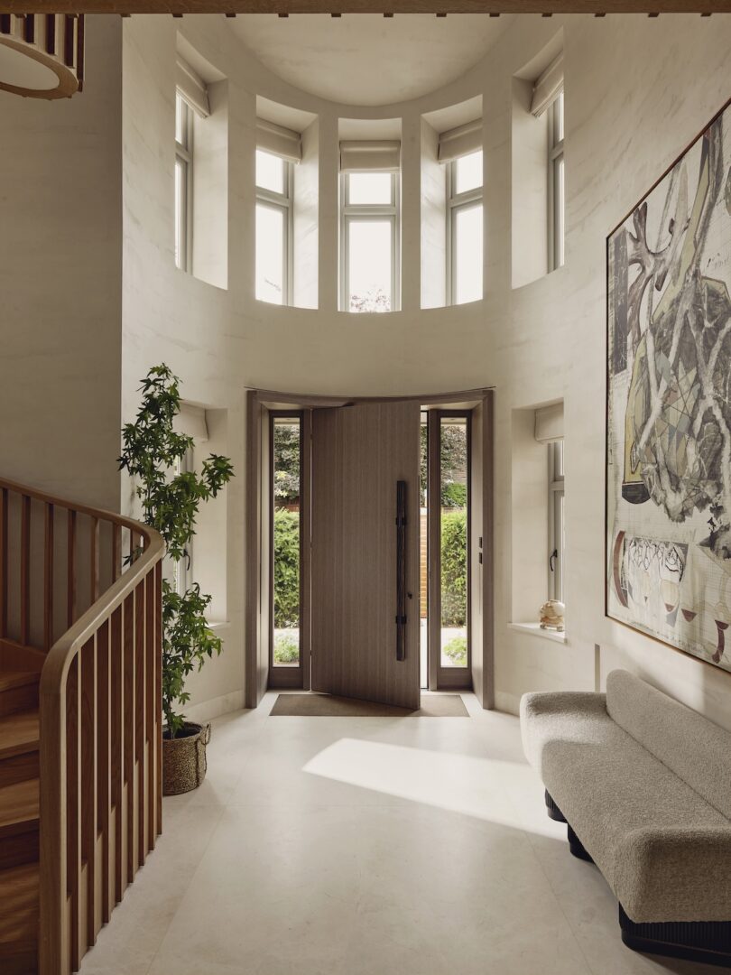 La entrada moderna cuenta con ventanas altas, una escalera de madera, plantas en macetas, un gran cuadro abstracto, un sofá curvo y puertas dobles que conducen al jardín.