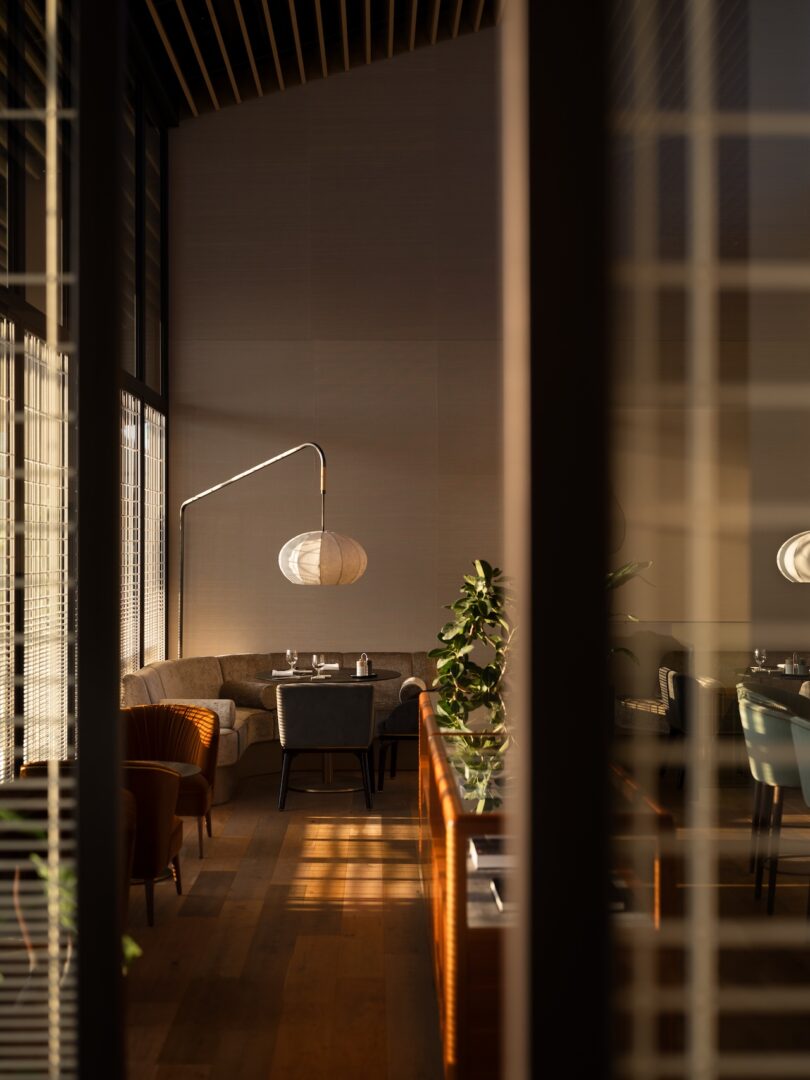 Interior de restaurante moderno con iluminación tenue, suelos de madera, asientos tapizados, lámparas de papel redondas y mesa de comedor visible a través de ventanas cerradas.