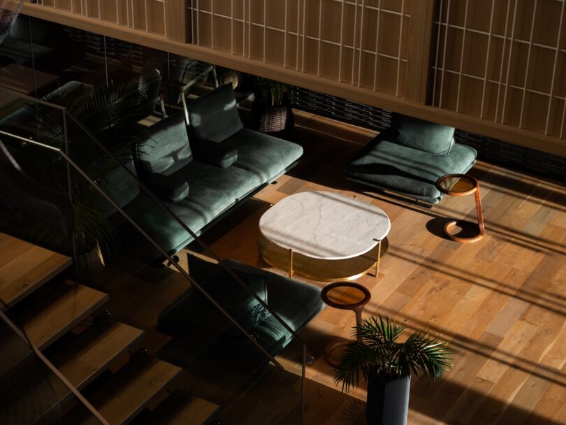 Salón moderno con sillas de terciopelo verde, mesa baja de mármol blanco, mesa auxiliar de madera y maceta sobre suelo de madera con luz natural.