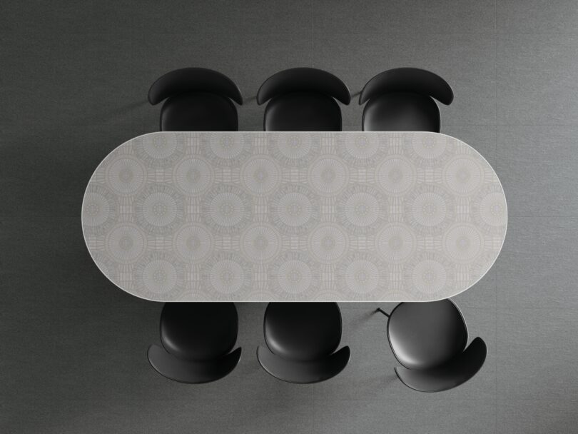 Vista de arriba hacia abajo de una mesa ovalada con una superficie estampada y seis sillas negras espaciadas uniformemente alrededor de la mesa sobre un piso gris