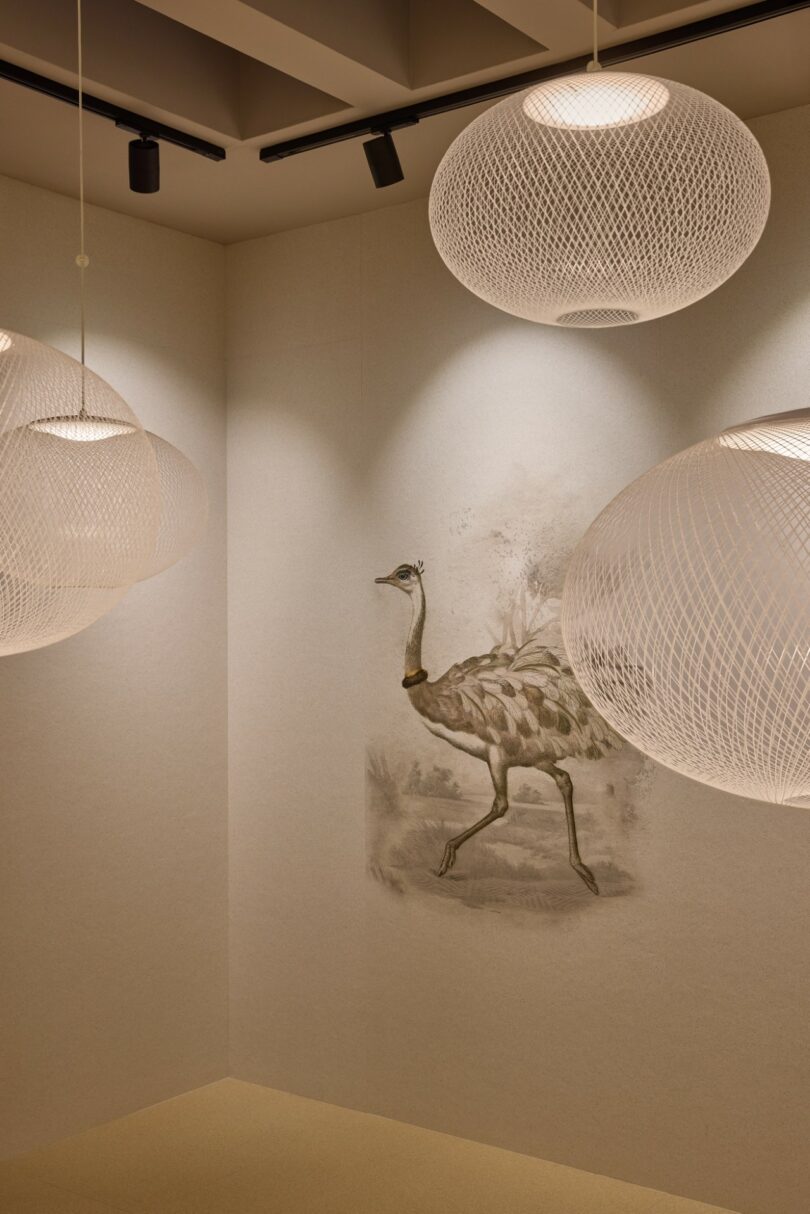 Tres candelabros esféricos cuelgan del techo, iluminando una habitación minimalista con un mural que representa un pájaro andante, posiblemente un avestruz o un emú, en tonos pastel.