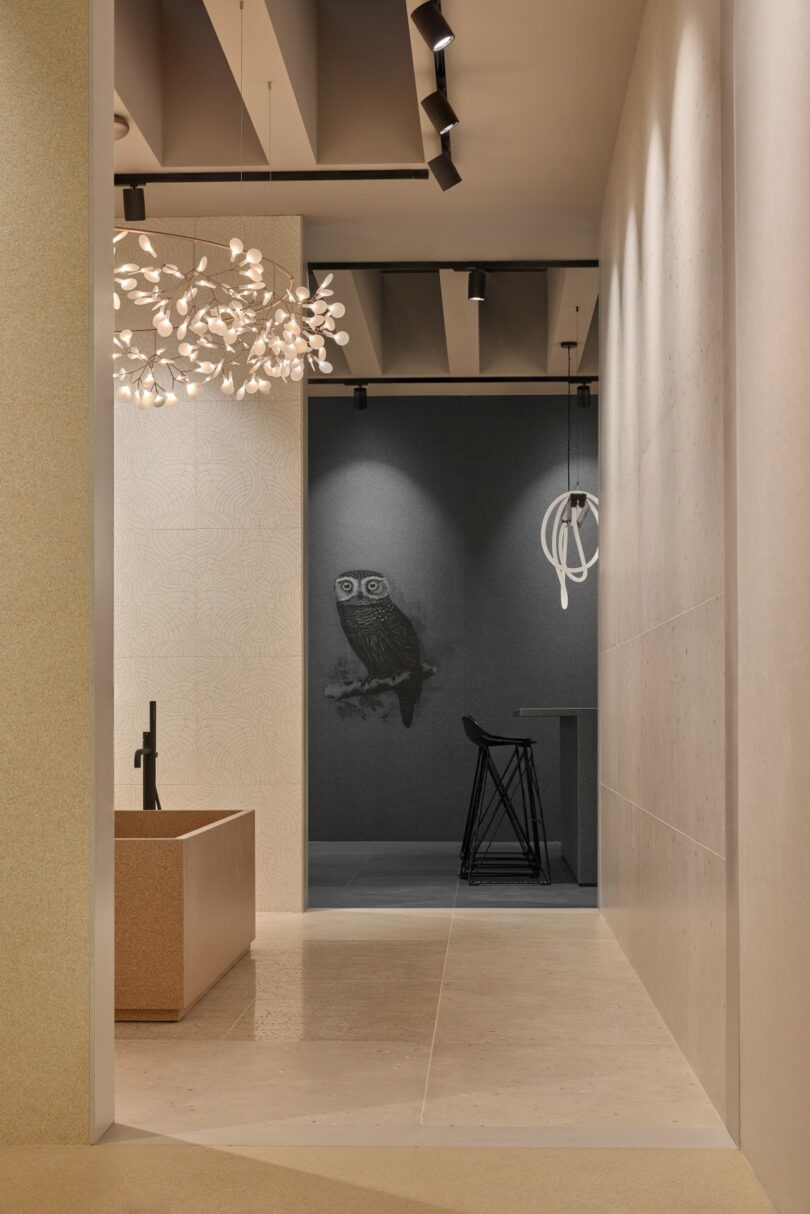 Pasillo moderno con suelo beige, lámpara de araña escultural, bañera independiente y paredes negras con ilustración de búho junto a taburetes negros y lámpara de araña