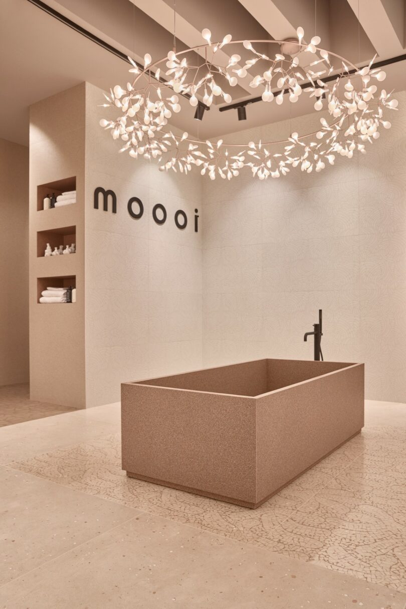 Cuarto de baño moderno y minimalista con bañera rectangular independiente, estante de pared con toallas y lámpara de araña decorativa; "mui" firmar en la pared