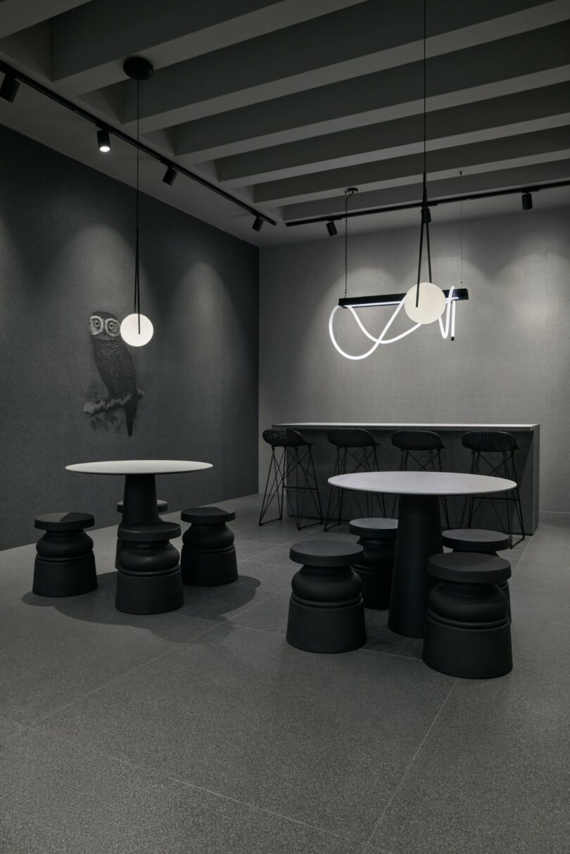 Interior moderno y minimalista de una cafetería con mesas redondas, taburetes negros, barra de bar con taburetes de estructura metálica y lámpara de araña geométrica; pared decorada con mural de búho