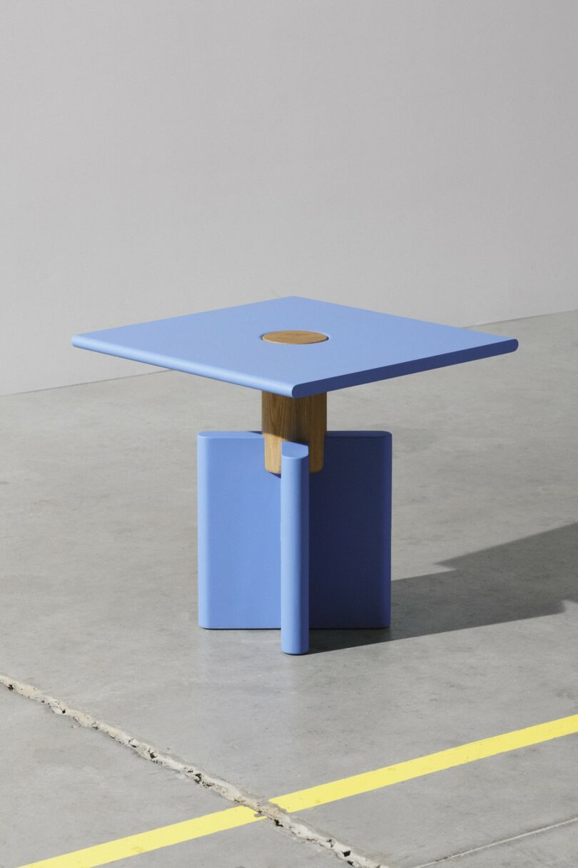 Una moderna mesa cuadrada azul con adornos de madera en el centro se alza sobre un suelo de hormigón pulido con una línea amarilla en el lateral