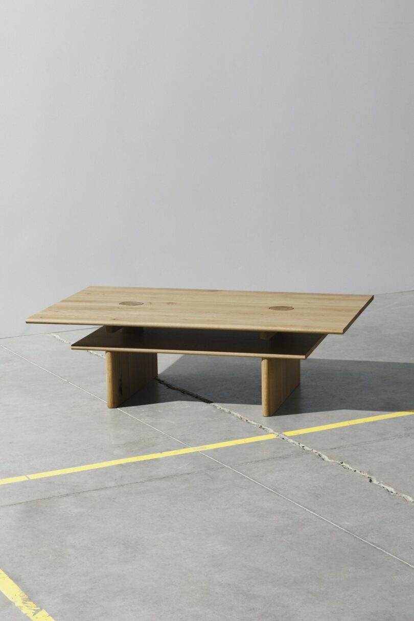 Una mesa de centro rectangular de madera con una parte superior plana y estantes inferiores se asienta sobre un piso de concreto gris con líneas de estacionamiento amarillas.