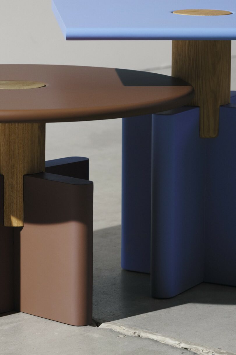 Dos mesas modernas con cantos redondeados y patas de madera; una mesa es marrón y la otra es azul, ambas sobre un piso de concreto