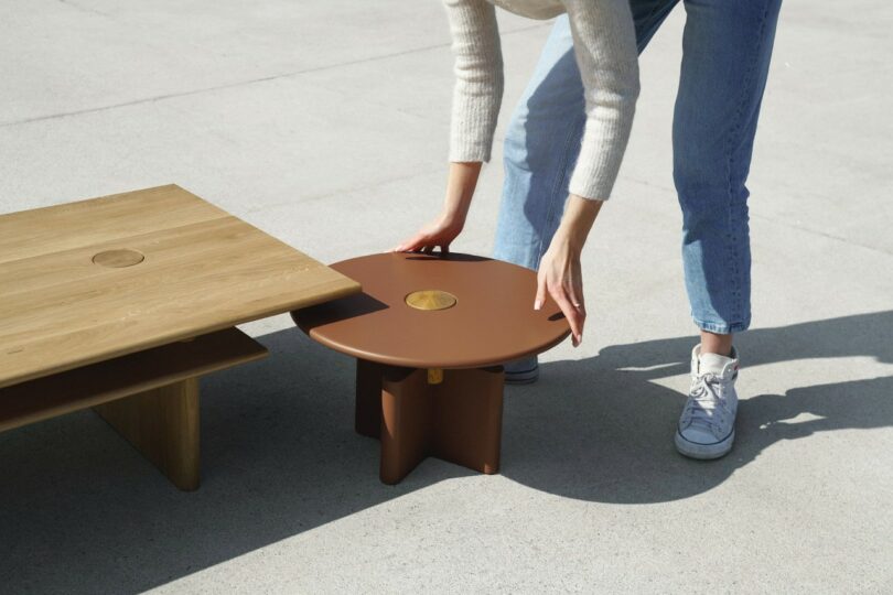 Un hombre con vaqueros y zapatillas de deporte ajusta una pequeña mesa redonda de color marrón junto a una mesa rectangular de madera más grande sobre una superficie lisa.