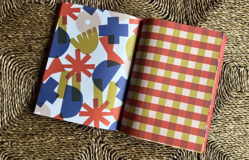 Un libro abierto de Design Milk Take 5 que muestra formas geométricas abstractas y coloridas en la página izquierda y un patrón de cuadros rojos, amarillos y blancos en la página derecha, colocados sobre una superficie tejida texturizada.