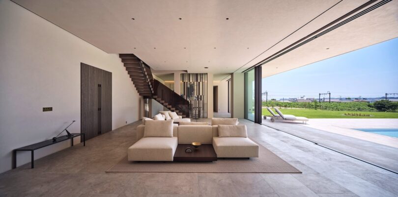 Salón moderno con sofá beige, mesa de centro de madera y pared abierta que da al patio, piscina y césped exterior con tumbonas y vistas al mar.