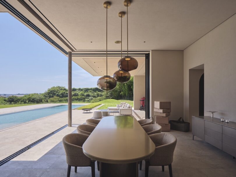 El moderno comedor cuenta con una mesa y sillas largas, una lámpara de araña y grandes puertas que se abren a las vistas a la piscina y al jardín; Varias cajas están apiladas contra la pared.