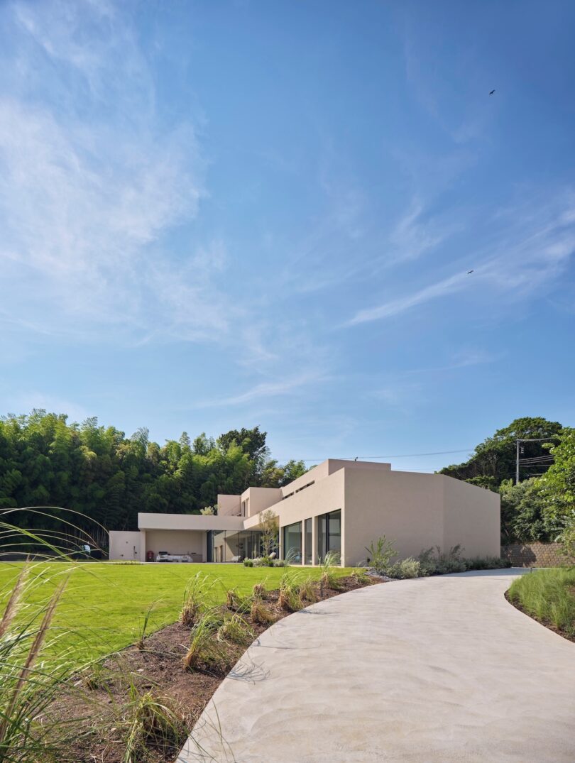 Una casa moderna de color beige con techo plano y grandes ventanales se encuentra junto a un sinuoso camino de concreto, rodeada de césped, plantas y árboles, bajo un cielo azul.