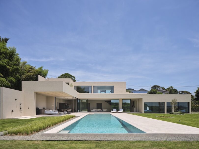 Casa moderna de dos pisos con techo plano, grandes ventanales de vidrio y una piscina rectangular en un patio trasero bellamente ajardinado bajo un cielo azul claro.
