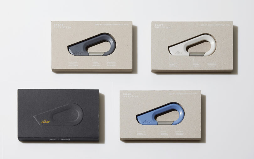 Cuatro objetos con forma de mosquetón en negro, blanco y azul están empaquetados en cajas minimalistas sobre una superficie blanca. Tres cajas son marrones y una es oscura con letras doradas.