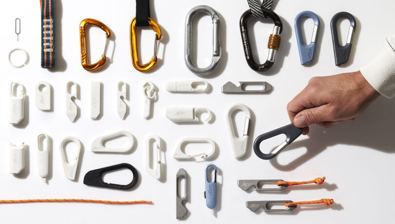 Sobre una superficie blanca se disponen mosquetones y clips de diversas formas, tamaños y colores. Una mano sostiene un objeto parecido a un mosquetón de color gris oscuro.