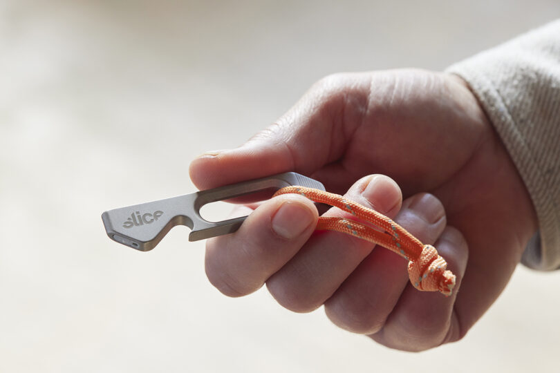 Una mano sostiene una pequeña multiherramienta de metal con una marca "pedazo" y una cuerda naranja adicional.