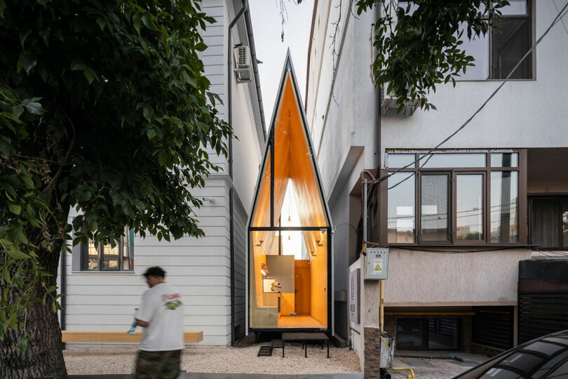 Una casa estrecha y moderna con un techo triangular afilado se asienta entre dos edificios más grandes; la entrada frontal de cristal emite un brillo cálido. Una persona pasa en primer plano.