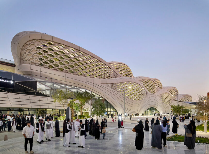 Al anochecer, la multitud se reúne frente a un edificio moderno con un techo ondulado en forma de celosía y grandes ventanales de vidrio. En primer plano se pueden ver árboles y zona ajardinada.