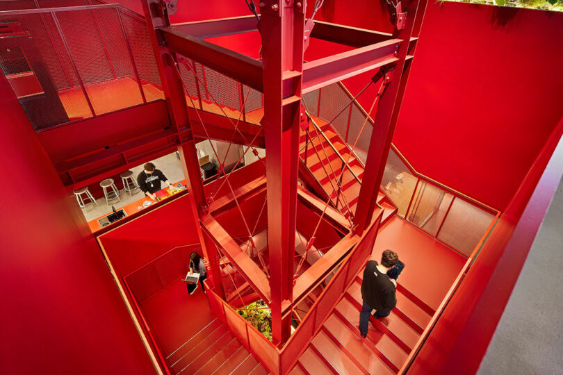 El luminoso interior de estilo industrial con paredes y escaleras rojas, con dos personas caminando por las escaleras y una persona sentada en el mostrador, presenta una escena dinámica perfecta para el foco de atención de Friday Five.