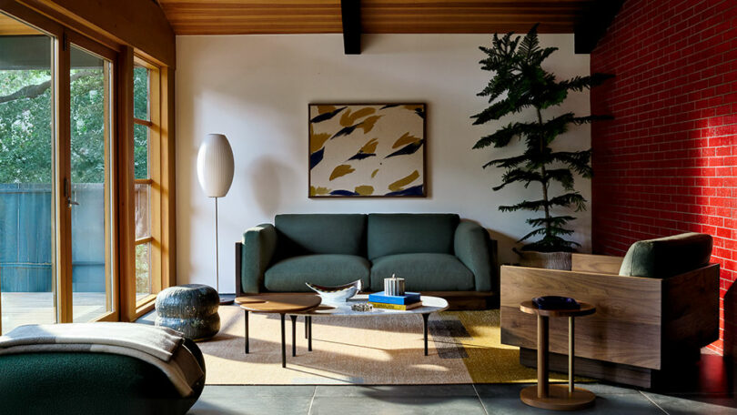 Salón moderno con sofá verde oscuro, sillón de madera, arte abstracto en la pared, plantas en macetas y grandes ventanales. Impresionante pared de ladrillo rojo.