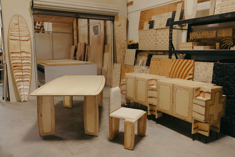El taller exhibe muebles de madera contrachapada sin terminar, incluidas mesas, sillas y gabinetes, así como tablones de madera y tablas de surf apoyadas contra las paredes.