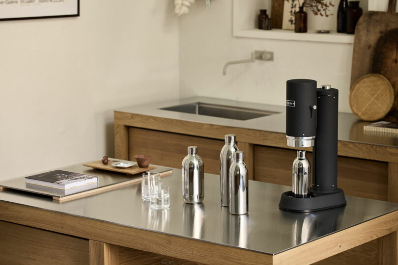 Una máquina de refrescos negra y tres botellas de metal se colocan sobre una encimera de metal en una cocina moderna con gabinetes de madera.