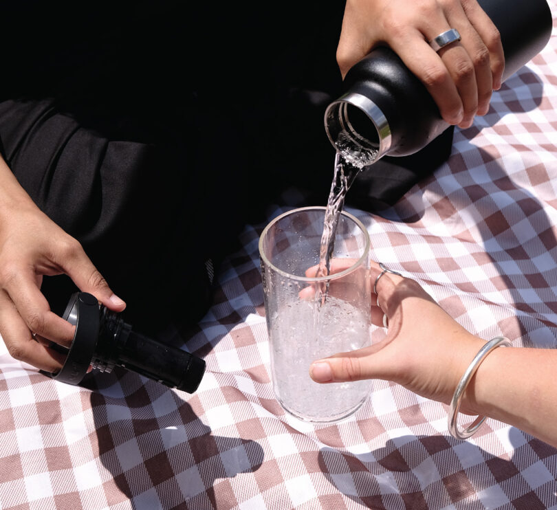 Dos personas se sientan sobre una manta de picnic a cuadros, una vierte agua de un termo negro en un vaso transparente sostenido por la otra