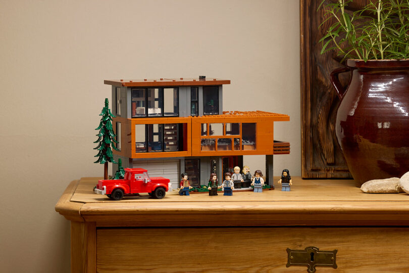Un modelo de casa moderna de Lego con un camión rojo, seis figuras de Lego y un árbol exhibido en una caja de madera junto a una gran maceta marrón con plantas verdes