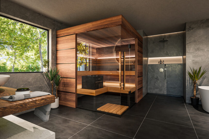 Cuarto de baño moderno con una gran sauna de madera acristalada, paredes y suelos de piedra gris, ducha a ras de suelo, plantas y ventana con vistas a la vegetación