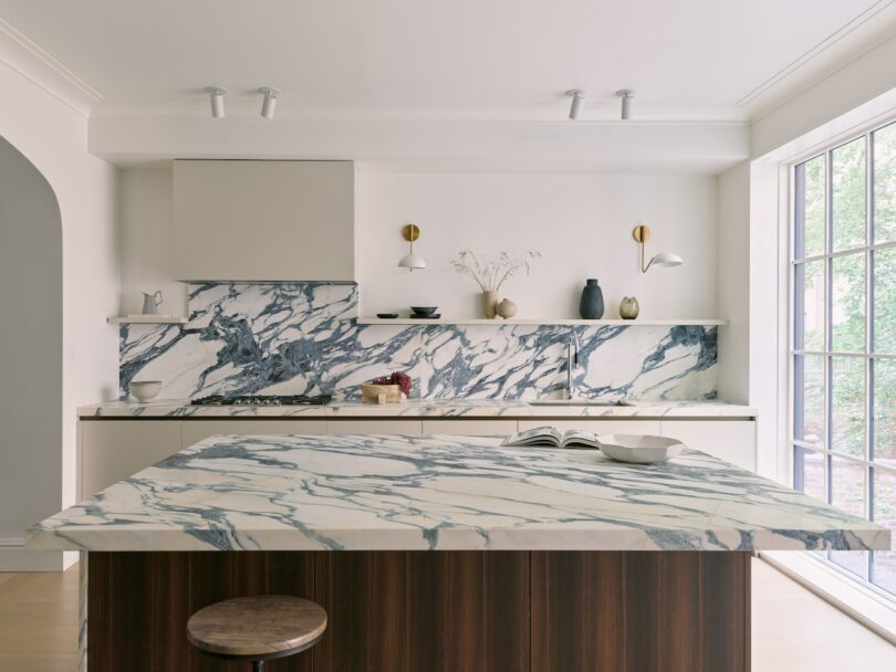 Modern kitchen with white and blue marble countertops and backsplash, wood island with a stool, open shelving, and large window letting in natural light—perfect for enjoying your morning coffee with skim milk.