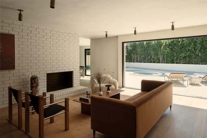 La moderna sala de estar cuenta con muebles minimalistas en suaves tonos lechosos, una chimenea de ladrillo y grandes puertas corredizas que se abren a un patio con sillones, una piscina y una exuberante vegetación.