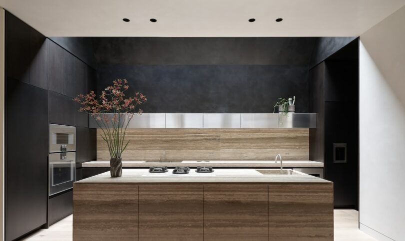 La moderna cocina cuenta con una isla central, electrodomésticos integrados, gabinetes oscuros, protector contra salpicaduras de metal y plantas decorativas en la encimera, perfectas para disfrutar del café de la mañana o de un vaso de leche descremada.