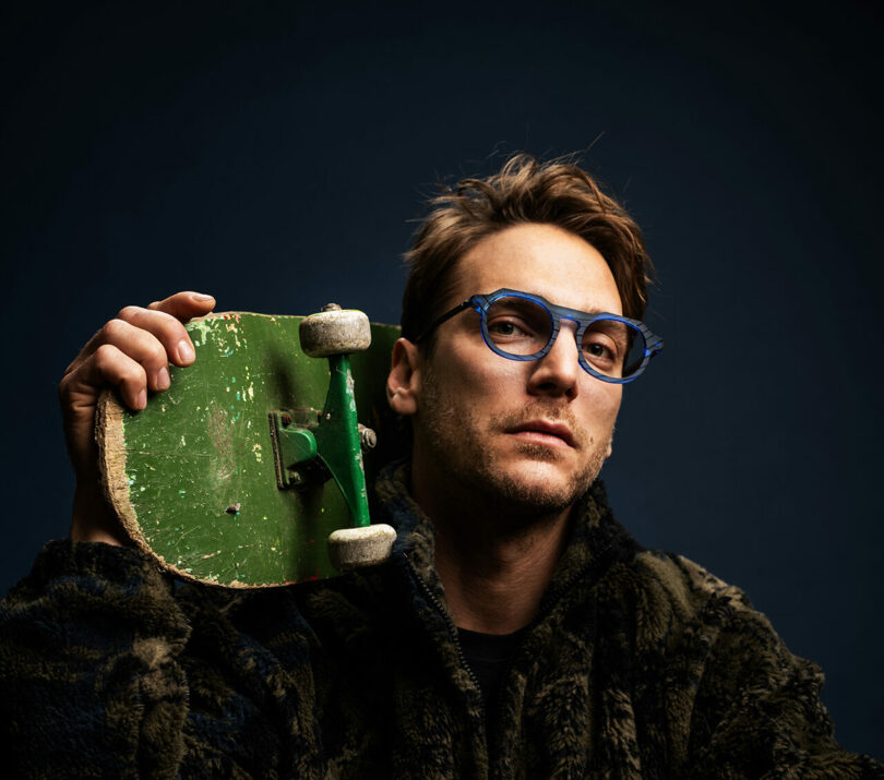 Un hombre que lleva las 10 mejores gafas azules de moda de 2025 y una chaqueta de textura oscura sostiene una patineta verde desgastada sobre sus hombros sobre un fondo oscuro.