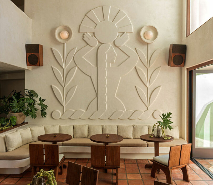 Esta habitación iluminada por el sol, centrada alrededor de un banco tapizado de color beige, una mesa de madera, sillas y plantas en macetas, está coronada por un mural tallado de una mujer con cabello largo sosteniendo un círculo, perfecto para su próximo Top 10 de lugares de inspiración para viajes para 2025.