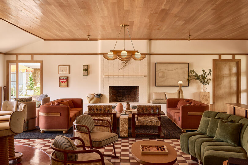 El espacioso salón cuenta con una chimenea central de ladrillo blanco, varios sillones y sofás estampados, un techo de madera, paredes de colores claros y obras de arte modernas en las paredes.