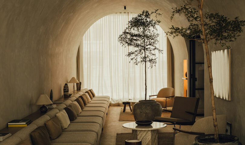 Salón moderno con techo abovedado, sofá beige, sillón, árbol en maceta, mesa de centro redonda y cortinas transparentes que van del suelo al techo que dejan entrar la luz natural.