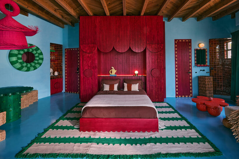 El dormitorio está inspirado en los 10 mejores viajes de 2025, con una cabecera de caoba, paredes azules, alfombras a rayas verdes y blancas y una decoración ecléctica con colores llamativos y patrones geométricos.