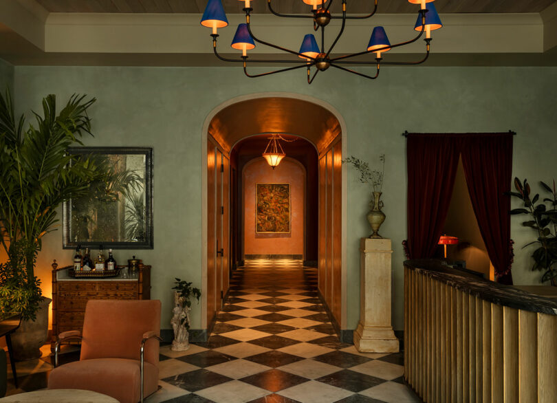 El vestíbulo del hotel, cálidamente iluminado, cuenta con pisos a cuadros, pasillos con arcos, muebles eclécticos, plantas de interior y una lámpara de araña con tonos azules.