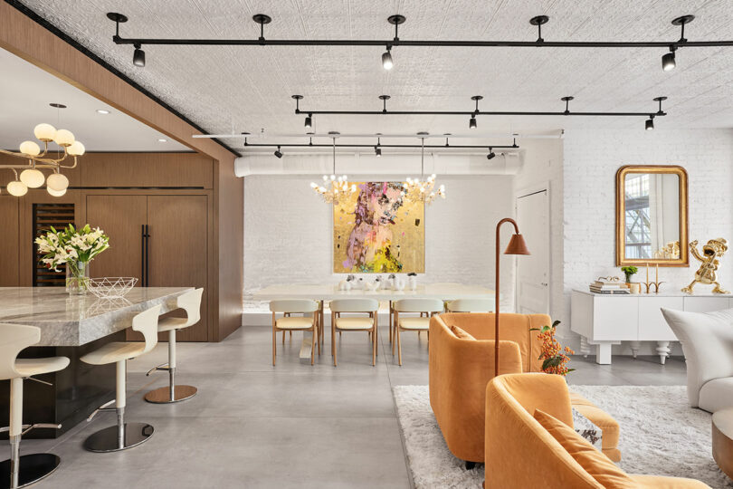 Sala de estar moderna y abierta con cocina, comedor y salón en tonos neutros, detalles dorados, muebles modernos y coloridas obras de arte en paredes de ladrillo blanco. 