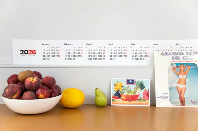 En la pared encima de la mesa cuelga un calendario para el año 2026 con un cuenco de melocotones, un limón, una pera, una postal en color y un disco de vinilo con una mujer en bañador.